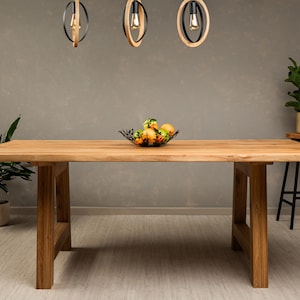 May include: A wooden dining table with a natural finish and a simple, rectangular design. The table has two trestle legs on each side and is set against a gray wall. A bowl of fruit sits on the table's surface.