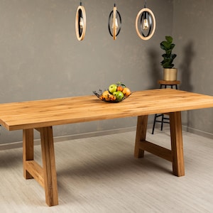 May include: A large, rectangular wooden dining table with a natural wood finish. The table has a simple, modern design with two A-frame legs on each side. The table is set with a bowl of fruit and is illuminated by three pendant lights.