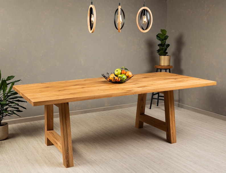 May include: A large, rectangular wooden dining table with a natural wood finish. The table has a simple, modern design with two A-frame legs on each side. The table is set with a bowl of fruit and is illuminated by three pendant lights.