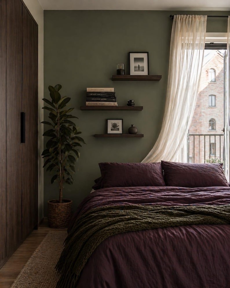 Puede incluir: Un dormitorio con una colcha y almohadas de color burdeos intenso, complementado con una manta de punto verde. La habitaci&oacute;n cuenta con una gran planta en maceta, estanter&iacute;as de madera oscura con libros y arte enmarcados, y una cortina blanca transparente. Las paredes est&aacute;n pintadas de verde apagado.