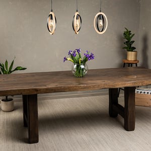 Puede incluir: Una gran mesa de comedor de madera marrón oscuro con borde natural. La mesa está sostenida por patas gruesas de color marrón oscuro. Un jarrón de cristal con flores moradas está sobre la mesa. Tres lámparas colgantes encima.