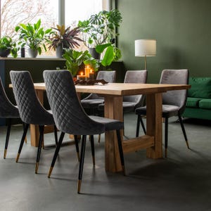 Può includere: Un tavolo da pranzo in legno con finitura naturale e gambe in metallo nero. Sei sedie in velluto grigio con dettagli dorati circondano il tavolo. Le sedie hanno un design trapuntato. Il tavolo è apparecchiato con candele e un pezzo decorativo.