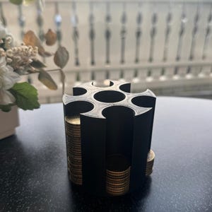 Könnte beinhalten: Ein schwarzer, zylindrischer Münzhalter mit sieben Fächern, die jeweils mit Stapeln goldfarbener Münzen gefüllt sind. Der Halter steht auf einer dunklen Oberfläche, mit einem verschwommenen Hintergrund aus Fenster und Blumen.