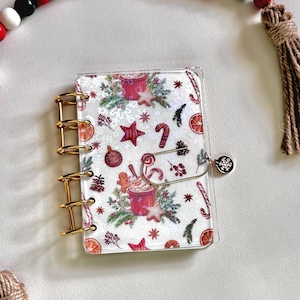 May include: A small, clear planner with gold-colored rings and a festive Christmas-themed cover. The cover features a pattern of red stars, candy canes, snowflakes, and coffee cups. A decorative tassel and beaded garland are in the background.