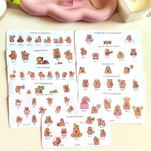 May include: Several sticker sheets featuring bear illustrations in various poses and activities. The sheets include phrases like "Cooking Up Greatness" and "Life's an Adventure." A pink tray with jewelry and a candle are also visible.