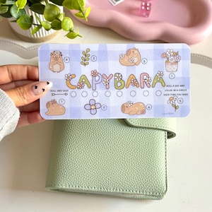 May include: A light blue and white patterned Capybara-themed savings tracker card. The card features illustrations of capybaras, flowers, and text that reads "CAPYBARA" and "ROLL AND SAVE". A light green leather wallet is in the foreground.