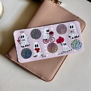 May include: A pink card featuring ghost illustrations with hearts and glitter circles, resting on a blush-colored wallet. The ghosts are depicted with various Valentine's Day themes, such as holding hearts and riding a bicycle. The card has the text "$2-$9" and "$75".