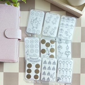 May include: A set of clear plastic savings challenge inserts featuring black line art illustrations of ducks, birthday cakes, balloons, and gift boxes. The cards include circular scratch-off areas, numerical values, and text such as Birthday Celebration, Birthday Balloons, Birthday Gifts, Birthday Wishes, Birthday Cake, and Birthday Surprise.