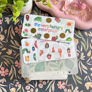 May include: Two rectangular savings challenge trackers featuring The Very Hungry Caterpillar theme. The top card has gold scratch-off circles and colorful illustrations of food and insects. The bottom card displays various food icons with text.