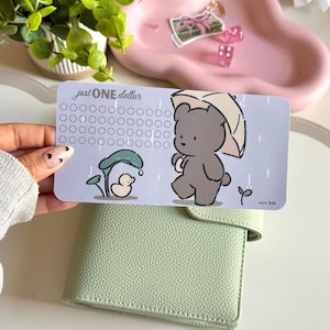 May include: A light blue savings tracker card with the text "just ONE dollar" and a cartoon bear with an umbrella. The card has circles for tracking savings and a duck under a leaf. A light green planner is in the foreground.