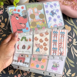May include: A clear plastic binder insert with three rows of pockets containing colorful savings challenge cards. The cards feature illustrations of Clifford the Big Red Dog, The Very Hungry Caterpillar, cookies, Thing 1 and 2, and The Cat in the Hat.