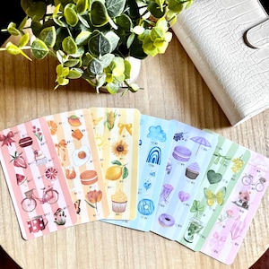 May include: A collection of colorful sticker sheets with various illustrations, including food, flowers, and rainbows. Each sheet features a different color scheme and striped background. A white planner and greenery are in the background.