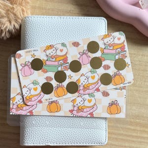 May include: Two rectangular savings challenge cards featuring a repeating pattern of sleeping cats on books, pumpkins, and pinecones. The top card has gold scratch-off circles, the text COZY FALL, and SAVE $50. The bottom card has the text $1-$10.