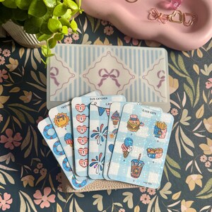 May include: A set of illustrated cards with a clear plastic case. The cards feature patriotic designs, including a sun, ice cream, and a burger. The case has a light blue and white striped pattern with bow accents.
