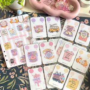 May include: A collection of rectangular cards featuring colorful illustrations and motivational text, including phrases like Badass Babe, Donut Worry, and Everything is going to be okay, arranged in a clear plastic sleeve alongside a sticker sheet.
