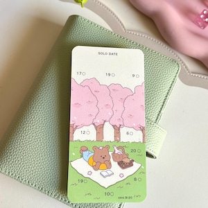 May include: A rectangular card with the text "SOLO DATE" at the top. The card features a cartoon bear on a picnic blanket under pink cherry blossom trees. Numbers are scattered around the image.