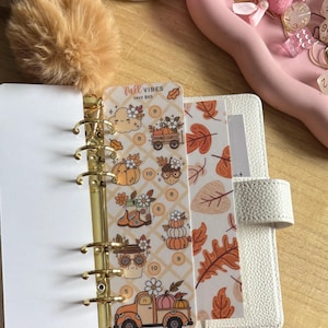 May include: A white six-ring binder containing a tracker card with the text "fall VIBES SAVE $65" and illustrations of pumpkins, a truck, and boots, alongside a second card featuring a repeating pattern of orange and brown autumn leaves.