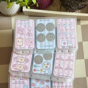 May include: A clear plastic binder insert with three vertical pockets containing printed savings challenge cards. The cards feature medical-themed illustrations like stethoscopes, bandages, and syringes in pink and blue stripes with silver scratch-off circles.