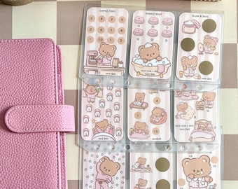 May include: A pink textured binder next to three clear plastic sheets containing nine individual savings challenge cards. Each card features brown bear illustrations, pastel striped backgrounds, and text including Coffee First, Bubble Bear, and Glow & Save, with various dollar amounts and circular scratch-off areas.