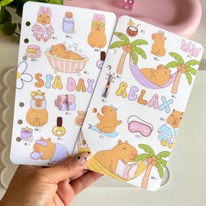 May include: Two planner sticker sheets featuring capybara illustrations in spa and relaxation settings. One sheet says "SPA DAY" and the other says "RELAX". The stickers include bath scenes, hammocks, and spa accessories.
