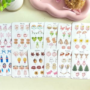 May include: A collection of twelve sticker sheets, each featuring themed illustrations for different months. Designs include seasonal elements like snowflakes, rainbows, flowers, and pumpkins. The stickers are arranged on a light-colored surface.