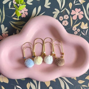 May include: Four large gold-toned metal paper clips, each adorned with a small, soft pom-pom in light blue, pale yellow, white, or brown. Each clip features small metallic beads and a single colored accent bead near the base of the pom-pom.
