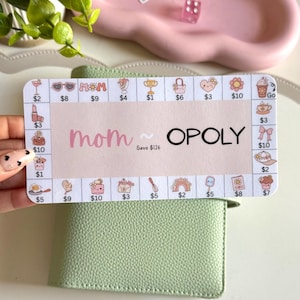May include: A light pink and white Mom-opoly game card with various icons and dollar amounts. The card is held over a light green wallet. The text "mom OPOLY" is displayed on the card.
