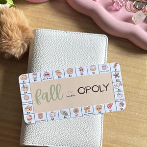 May include: A white textured leather-like planner cover with a rectangular game card titled Fall-Opoly. The card features a beige center with the text fall ~ OPOLY Save $100+ and a border of small autumn-themed icons with dollar amounts.