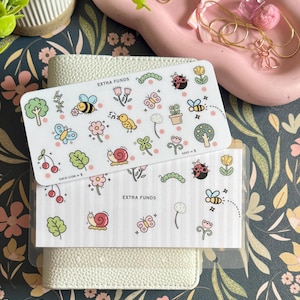 May include: Two rectangular white paper trackers labeled EXTRA FUNDS featuring colorful illustrations of nature elements including bees, butterflies, snails, ladybugs, flowers, trees, and cherries. The trackers are stacked on a white textured binder.