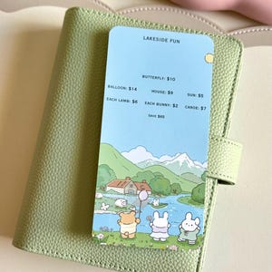 May include: A light green leather planner with a card featuring a lakeside scene with cartoon animals. The card has the text "LAKESIDE FUN" and prices for various items like balloons, houses, and bunnies. The planner is closed with a strap.