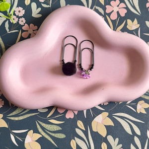 May include: Two black metal paper clips decorated with beads. One paper clip features a round, fuzzy black pom-pom, while the other is adorned with small black beads and a cluster of translucent, light purple flower-shaped charms.