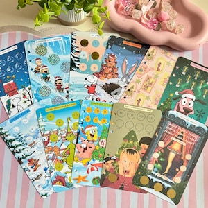 May include: A collection of ten rectangular paper savings trackers featuring various cartoon characters and holiday themes, including Snoopy, Bugs Bunny, SpongeBob, and Home Alone, with circular scratch-off areas and pre-punched holes for binder storage.