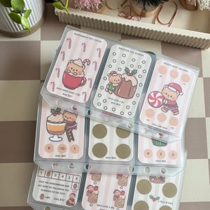 May include: A clear plastic binder page containing nine rectangular savings challenge cards with holiday-themed illustrations of bears, candy canes, and desserts. Each card features text, numerical values, and circular spots for tracking financial progress.