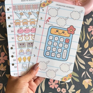 May include: Two white punched planner inserts with school-themed graphics. One sheet features a Back to School savings tracker with icons for pencils, buses, and apples. The other sheet, titled Crunching Numbers, displays a blue calculator graphic.
