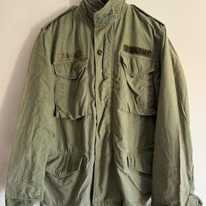 Può includere: Una giacca militare verde con una toppa dell'esercito americano sulla manica destra e una toppa "GENCK" sulla manica sinistra. La giacca ha una chiusura con cerniera e bottoni e diverse tasche.