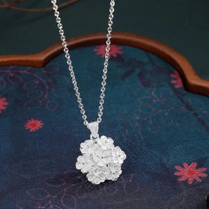 May include: A silver necklace featuring a cluster of small, detailed flower pendants. The pendant is suspended from a delicate chain. The background is a dark blue fabric with floral patterns and a wooden frame.