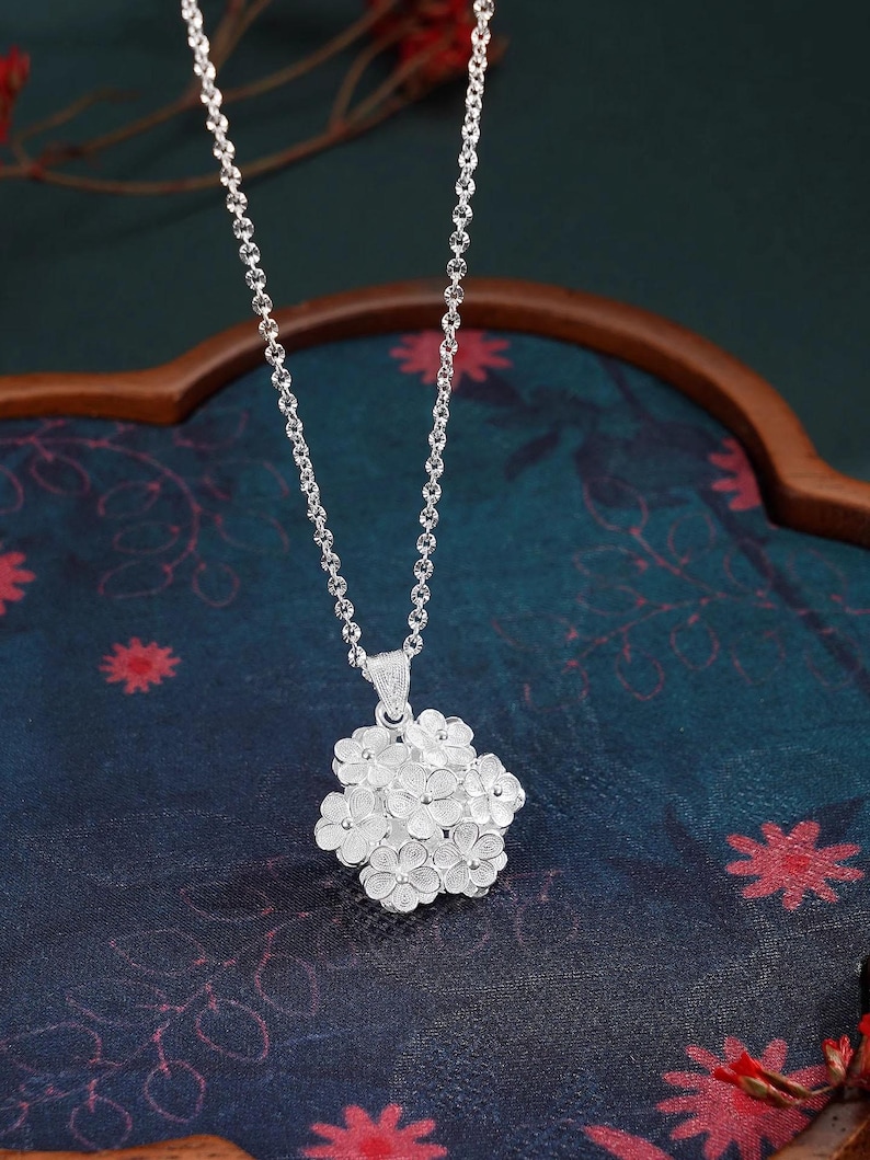 May include: A silver necklace featuring a cluster of small, detailed flower pendants. The pendant is suspended from a delicate chain. The background is a dark blue fabric with floral patterns and a wooden frame.