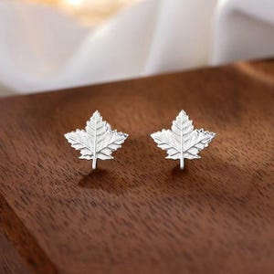 Puede incluir: Un par de pendientes de plata con forma de hoja de arce. Los pendientes están detallados y se muestran sobre una superficie de madera oscura.