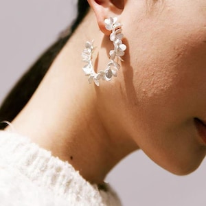 May include: Silver hoop earrings with a floral design. The earrings are made of silver and feature a series of small, delicate flower-shaped elements arranged around the hoop. The earrings are worn on the ear.