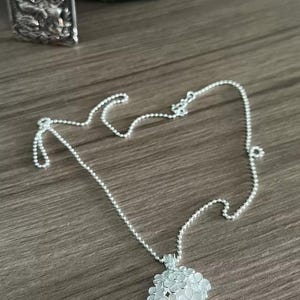 May include: A delicate silver necklace featuring a floral pendant. The pendant is a cluster of small, detailed flowers. The chain is a fine ball chain. The necklace is displayed on a wooden surface.