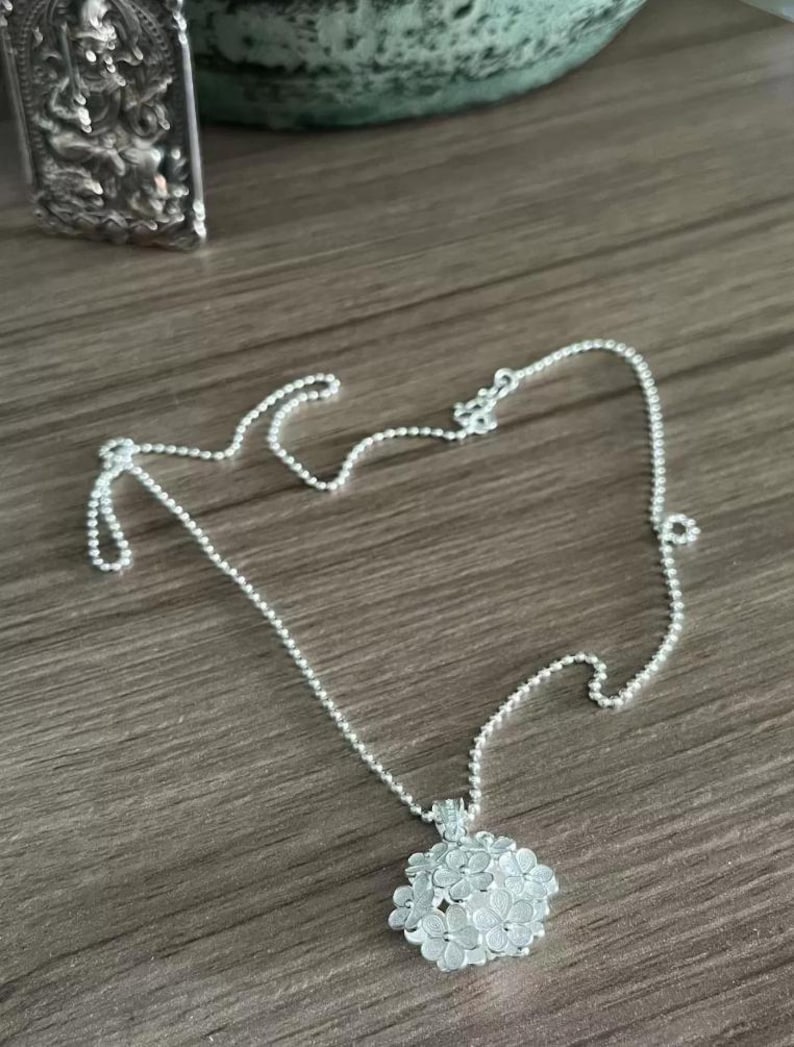 May include: A delicate silver necklace featuring a floral pendant. The pendant is a cluster of small, detailed flowers. The chain is a fine ball chain. The necklace is displayed on a wooden surface.