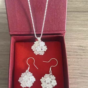 May include: A silver jewelry set featuring a necklace and a pair of earrings, presented in a red box. The necklace has a delicate chain and a floral pendant. The earrings also have a floral design. The jewelry is made of a shiny, reflective metal.