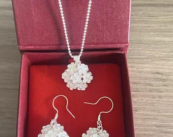 May include: A silver jewelry set featuring a necklace and a pair of earrings, presented in a red box. The necklace has a delicate chain and a floral pendant. The earrings also have a floral design. The jewelry is made of a shiny, reflective metal.