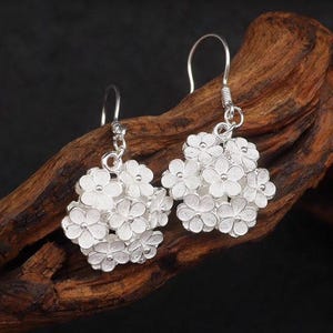 May include: A pair of silver earrings featuring clusters of small, detailed flowers. The earrings have a hook closure and are displayed against a dark background. The floral design is intricate and delicate, showcasing a classic jewelry style.