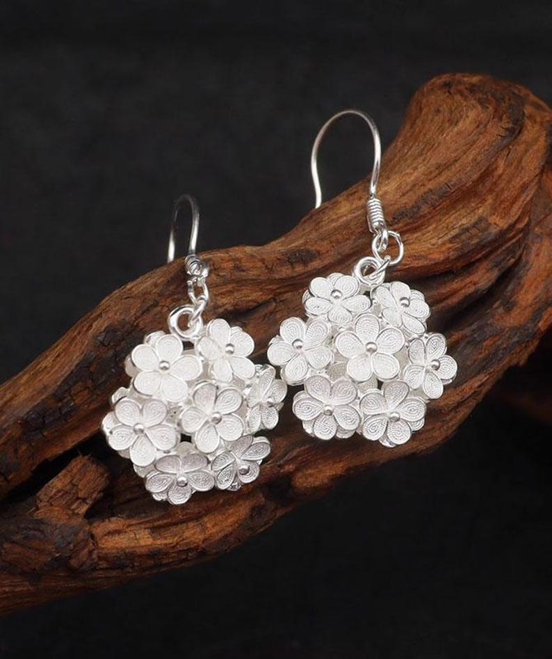 May include: A pair of silver earrings featuring clusters of small, detailed flowers. The earrings have a hook closure and are displayed against a dark background. The floral design is intricate and delicate, showcasing a classic jewelry style.
