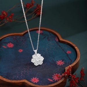 May include: A silver necklace with a floral pendant. The pendant is a cluster of small, detailed flowers. The necklace hangs on a delicate chain. The background is a dark blue surface with red floral accents.