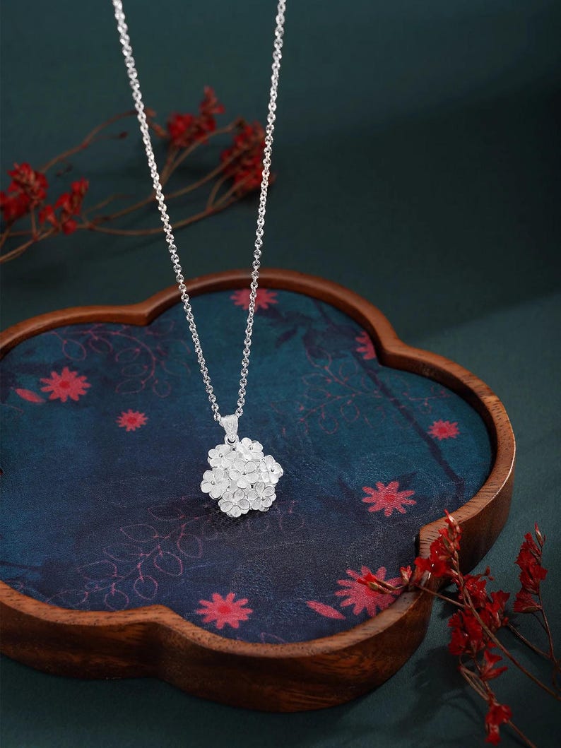 May include: A silver necklace with a floral pendant. The pendant is a cluster of small, detailed flowers. The necklace hangs on a delicate chain. The background is a dark blue surface with red floral accents.