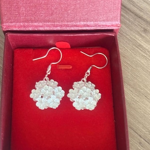 May include: A pair of silver-colored floral earrings displayed in a red jewelry box. The earrings feature a cluster of small, detailed flowers, hanging from a simple hook. The box is open, revealing a plush red interior.