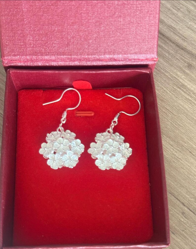 May include: A pair of silver-colored floral earrings displayed in a red jewelry box. The earrings feature a cluster of small, detailed flowers, hanging from a simple hook. The box is open, revealing a plush red interior.