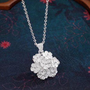 May include: A silver necklace featuring a cluster of small, detailed flower pendants. The pendant is suspended from a delicate chain. The necklace is displayed against a dark, patterned background.